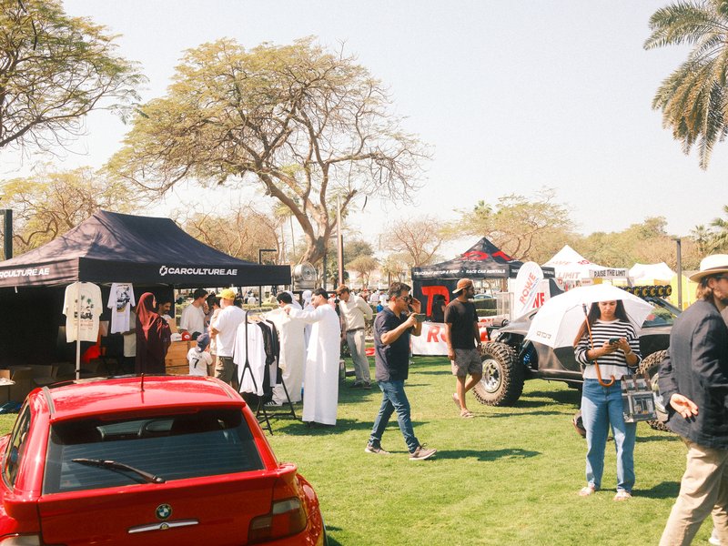 A vibrant outdoor market scene with tents, people browsing merchandise, and a red car in the foreground.