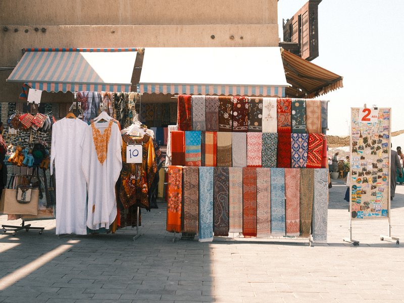 A vibrant outdoor market stall displaying colorful fabrics and traditional garments under bright sunlight.