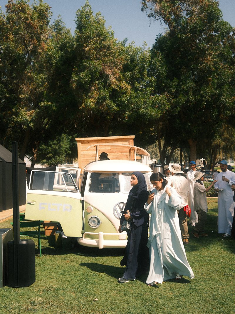 Two women pose in front of a vintage Volkswagen van at an outdoor event with trees in the background.