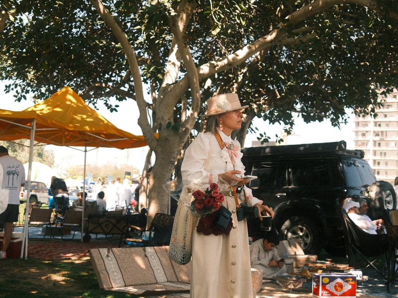 A man in a white outfit stands under a large tree at an outdoor event, with tents and people in the background.