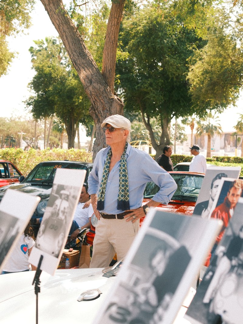 A man stands with hands on hips, surrounded by classic car photographs under a sunny tree.