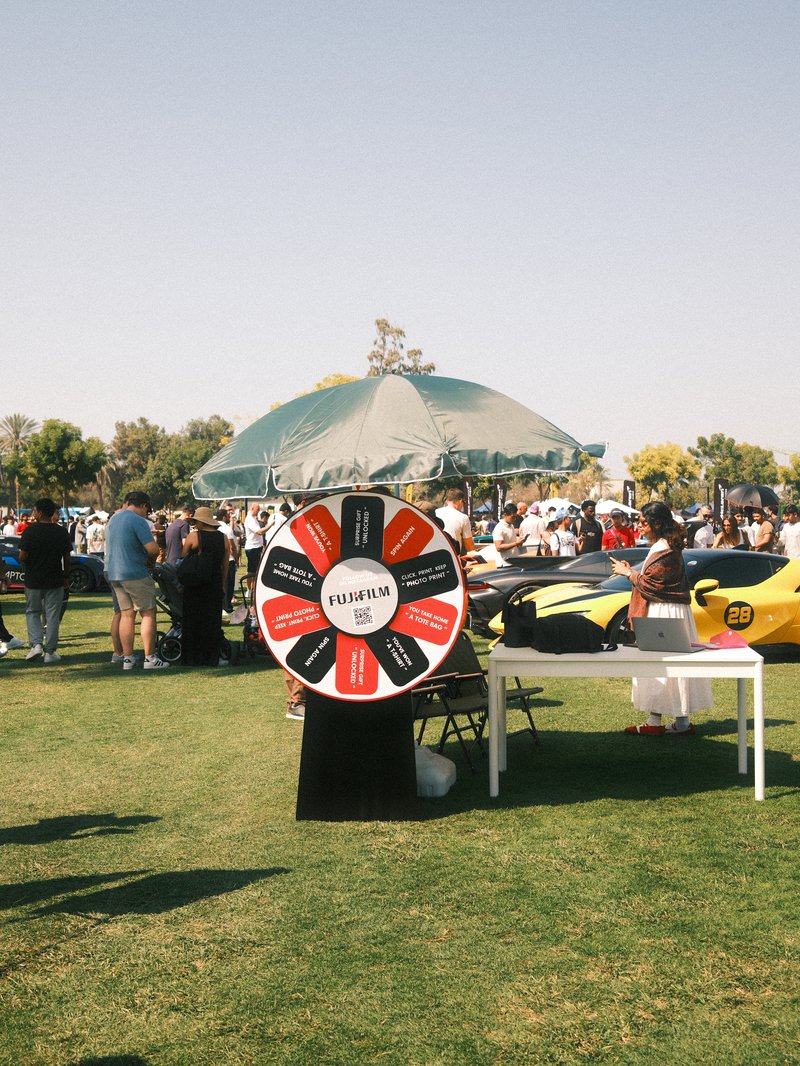 A colorful prize wheel under a green umbrella at an outdoor event with cars in the background.