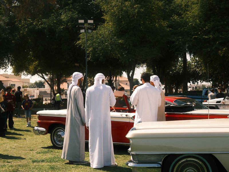 Three men in traditional attire observing classic cars in a park setting with trees.