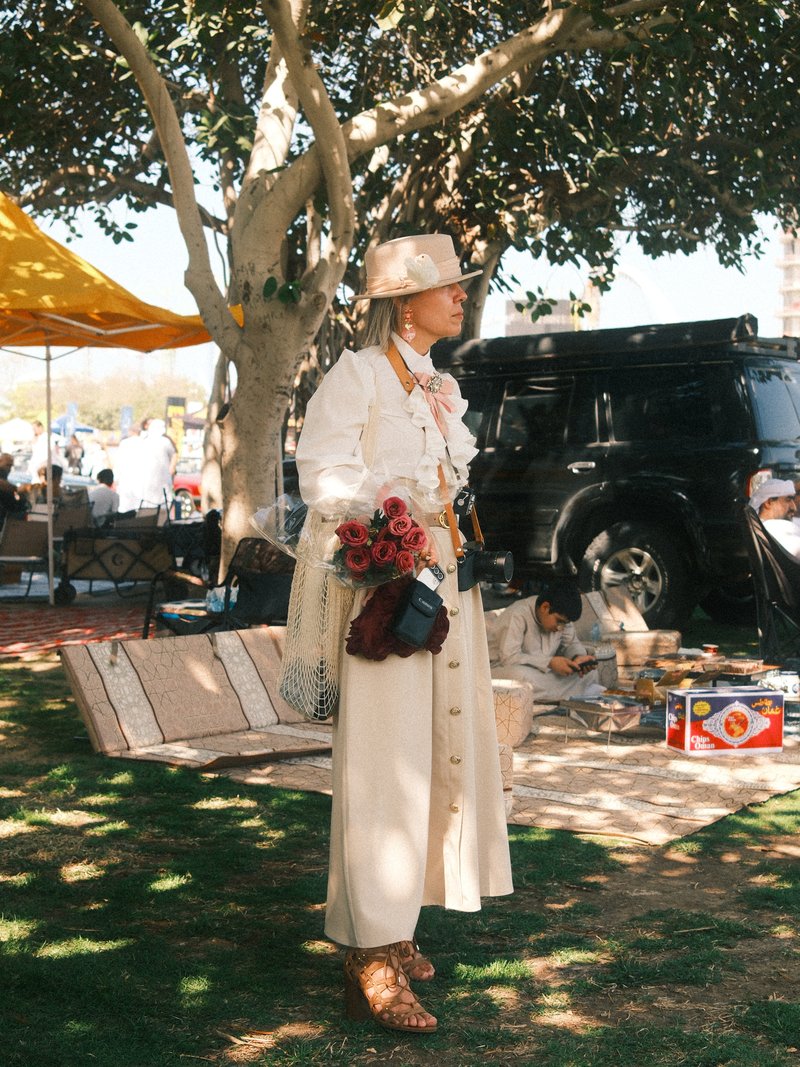 A stylish woman in a white outfit stands under a tree, holding flowers, with a black vehicle in the background.