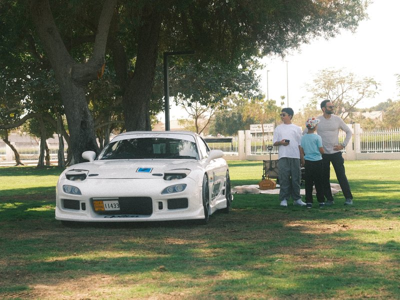 A white sports car parked on grass with three people standing nearby in a park.