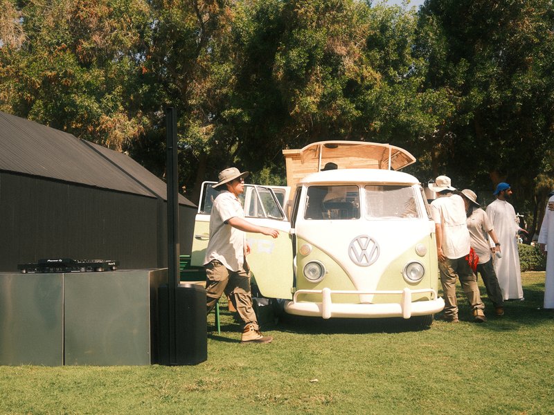 A vintage Volkswagen van parked on grass with people interacting around it in a sunny outdoor setting.