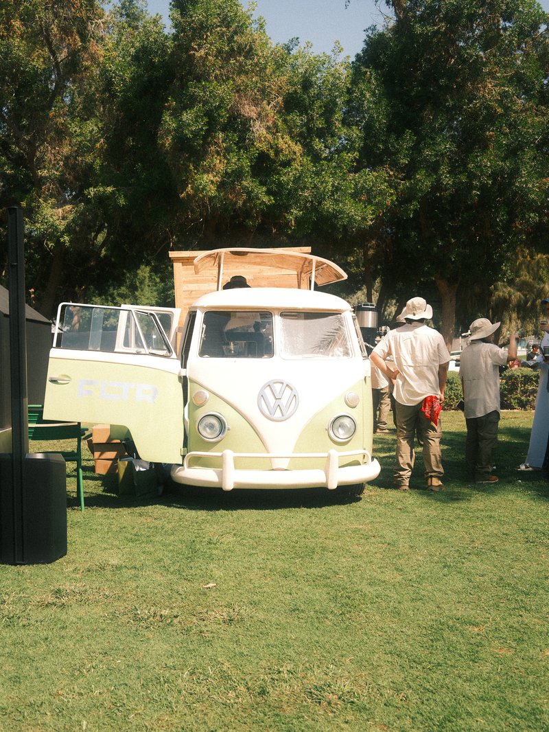 A vintage Volkswagen van parked on grass with people in hats nearby under trees.