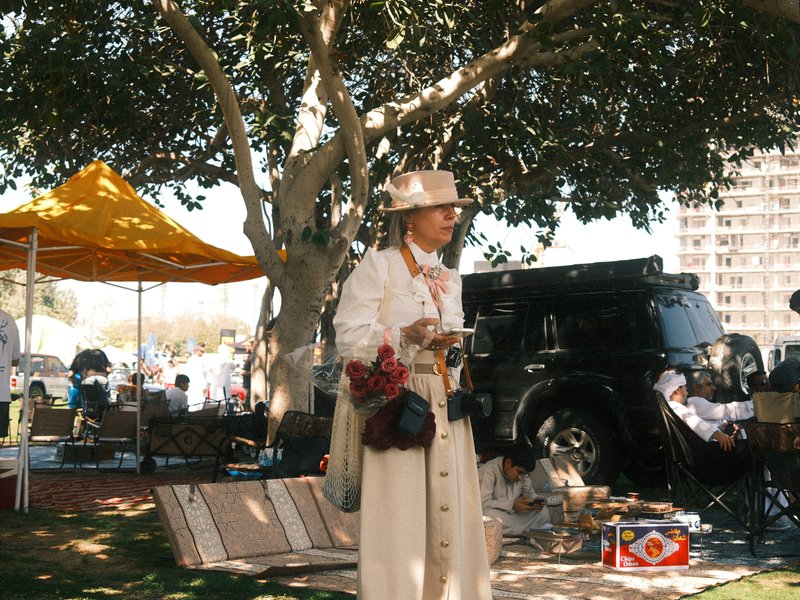 A person in a white coat and hat stands under a large tree at an outdoor market with tents.