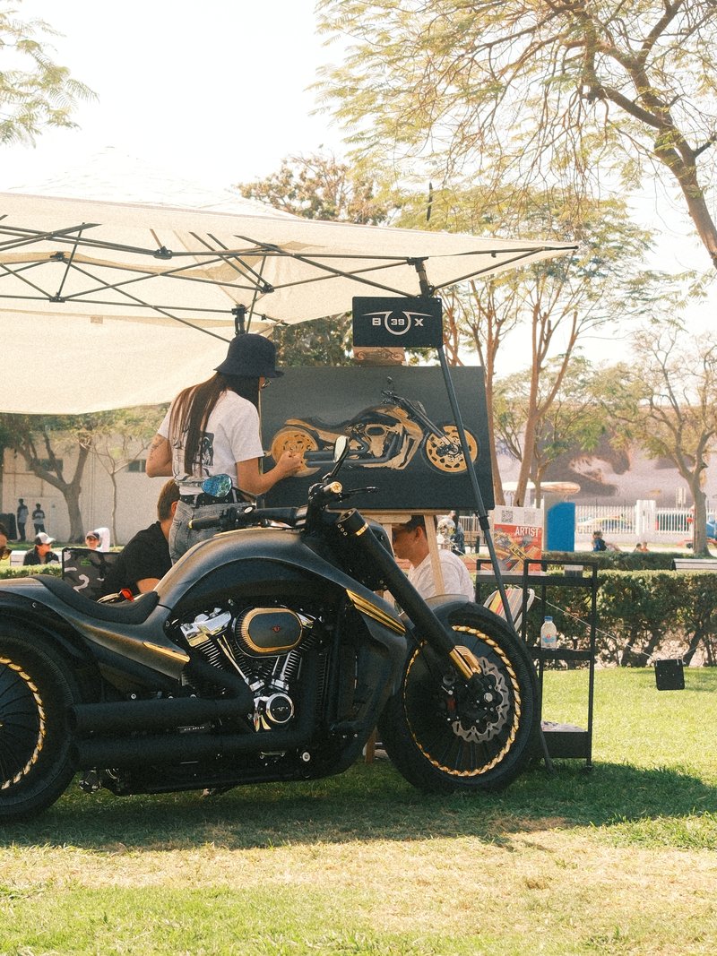 A custom motorcycle parked under a canopy with people and artwork in the background.