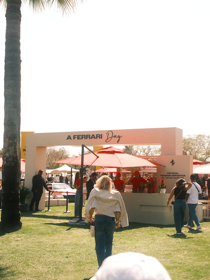 A crowd at a Ferrari Day event with a white booth and red umbrellas under bright sunlight.