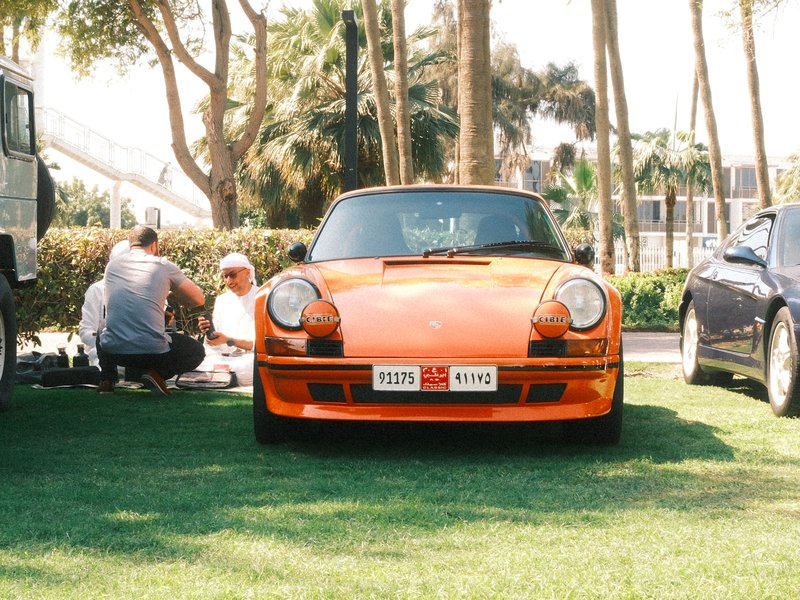 An orange vintage car parked on grass with people gathered nearby in a sunny outdoor setting.
