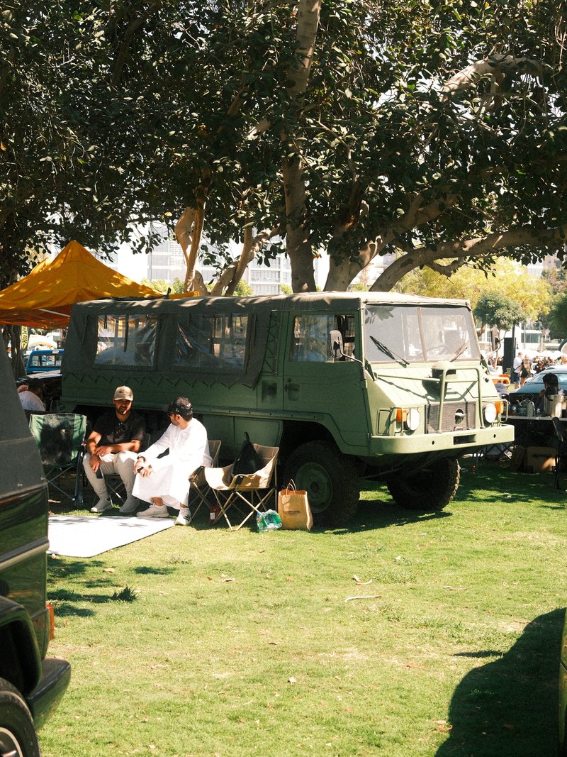 Two people relaxing beside a green military vehicle under a tree in a grassy area.