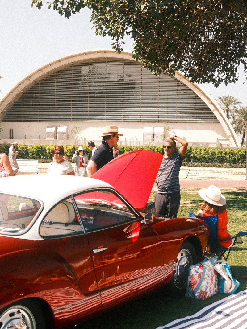 A classic red car parked in front of a modern building with people socializing in a sunny outdoor setting.