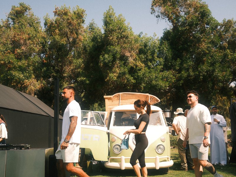 A vintage van in a lively outdoor setting with people walking around, surrounded by trees.