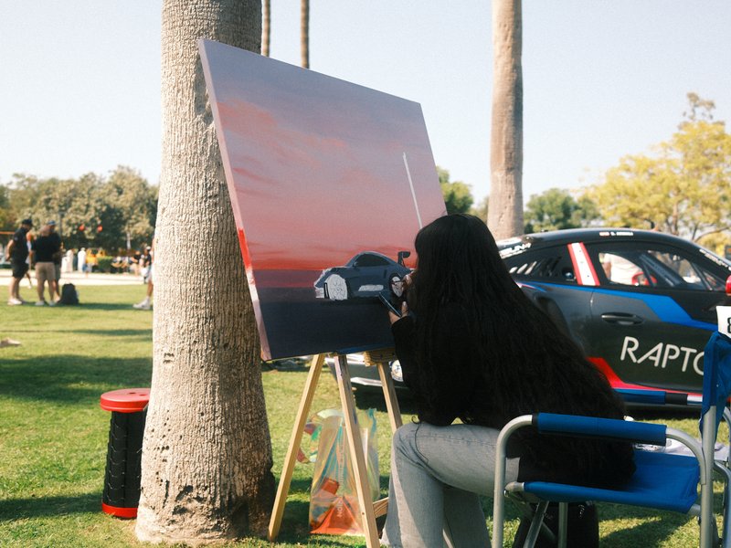 An artist painting a sunset scene on a canvas in a park with palm trees and cars in the background.