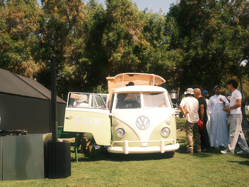 A vintage yellow Volkswagen van parked on grass with people gathered around it in a sunny outdoor setting.