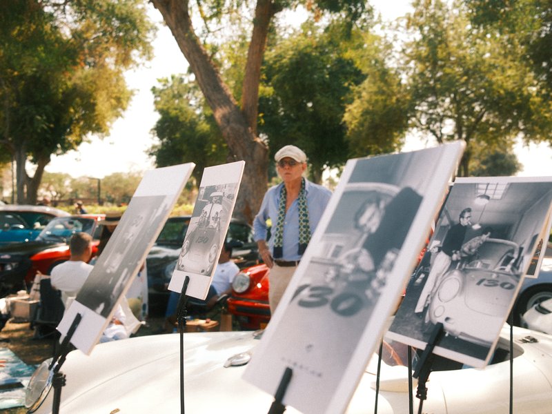 A man stands behind displayed photographs at an outdoor car show, with colorful cars in the background.