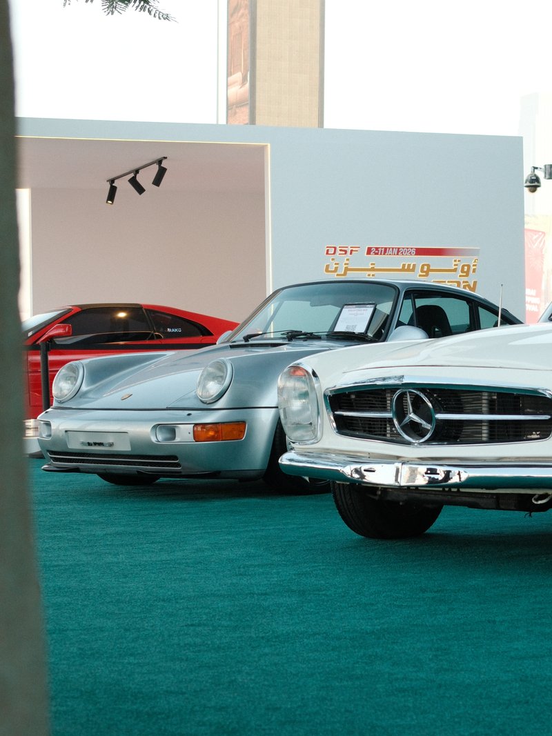 Two classic cars displayed at an exhibition, one silver and one red, on a green carpet.