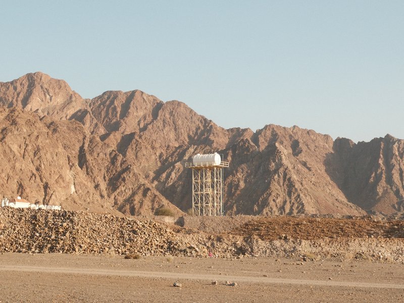 A water tower stands against a backdrop of rugged mountains in a desert landscape.