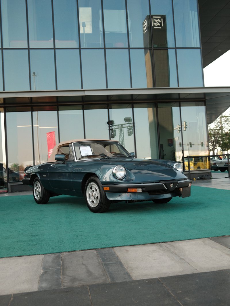 A vintage green convertible car parked on a green carpet in front of a modern glass building.