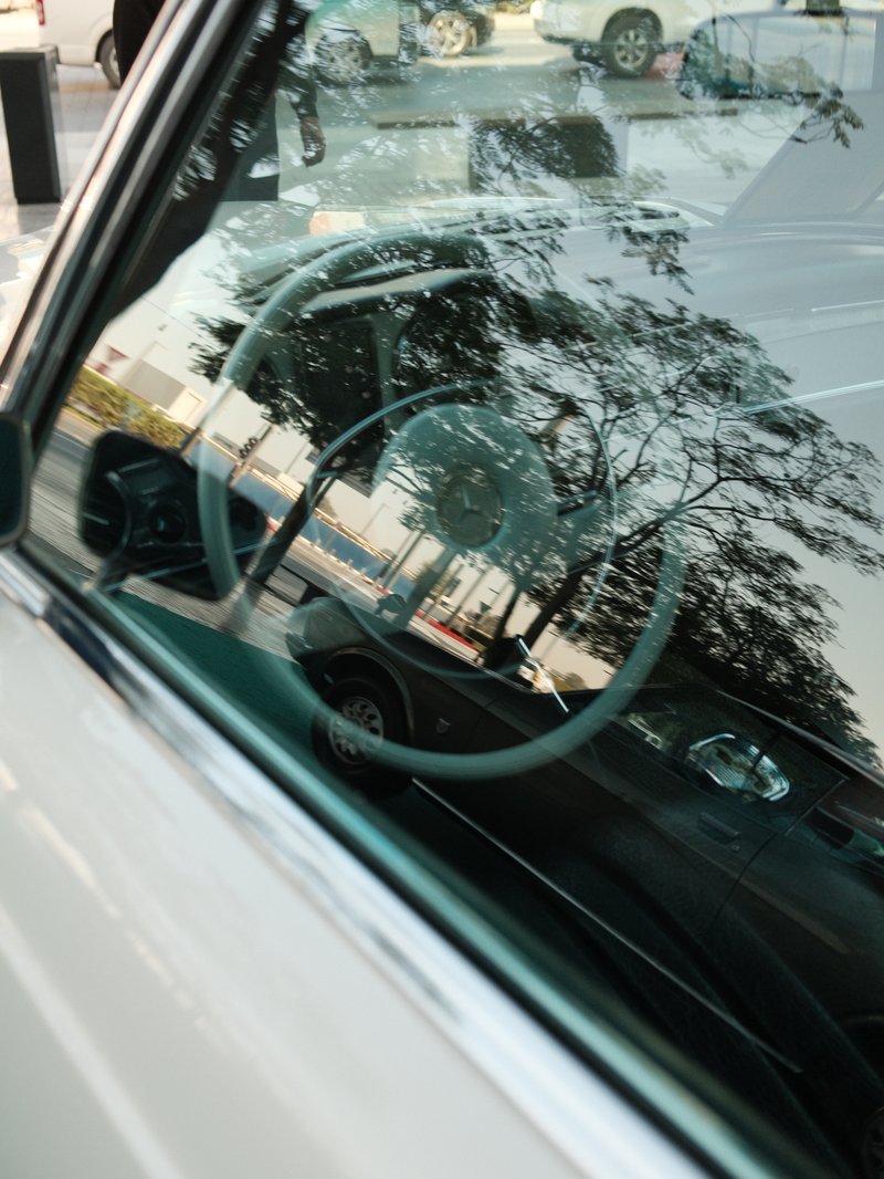 Close-up of a car's interior with a steering wheel reflected in the window and trees outside.