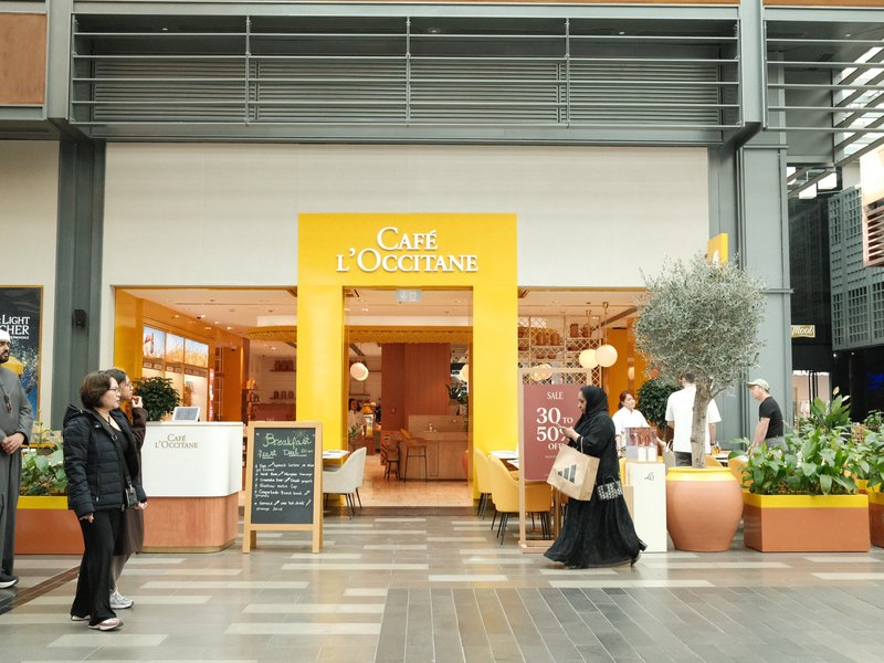 A café entrance in a shopping mall with people walking by and greenery in pots.