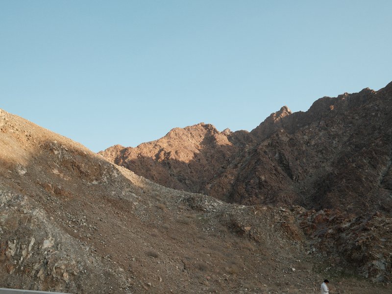 A rocky mountain landscape under a clear blue sky with warm sunlight illuminating the peaks.