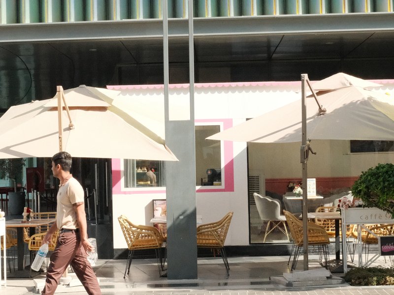 A man walks past a cafe with outdoor seating and large umbrellas in a modern urban setting.