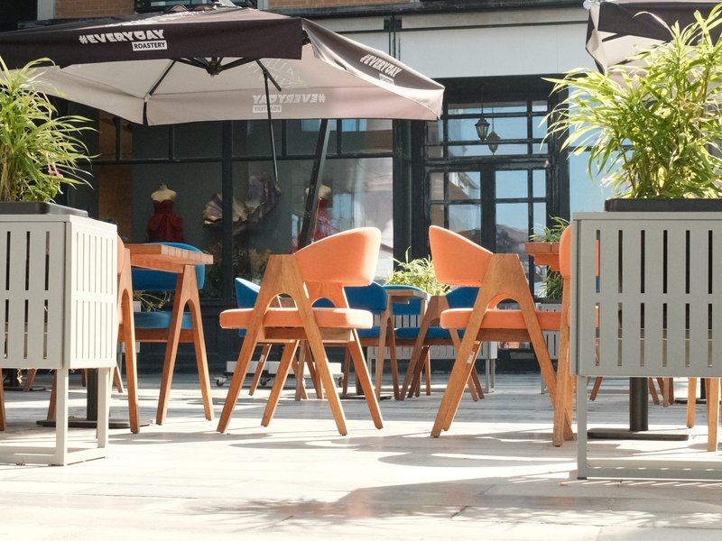 A sunny outdoor cafe scene with wooden chairs and umbrellas, surrounded by potted plants.