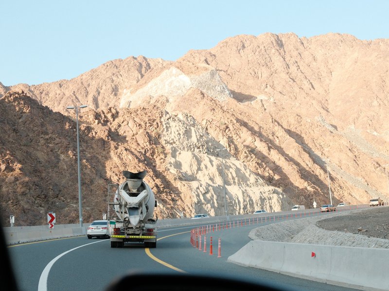 A cement mixer truck driving on a winding road with rocky mountains in the background.