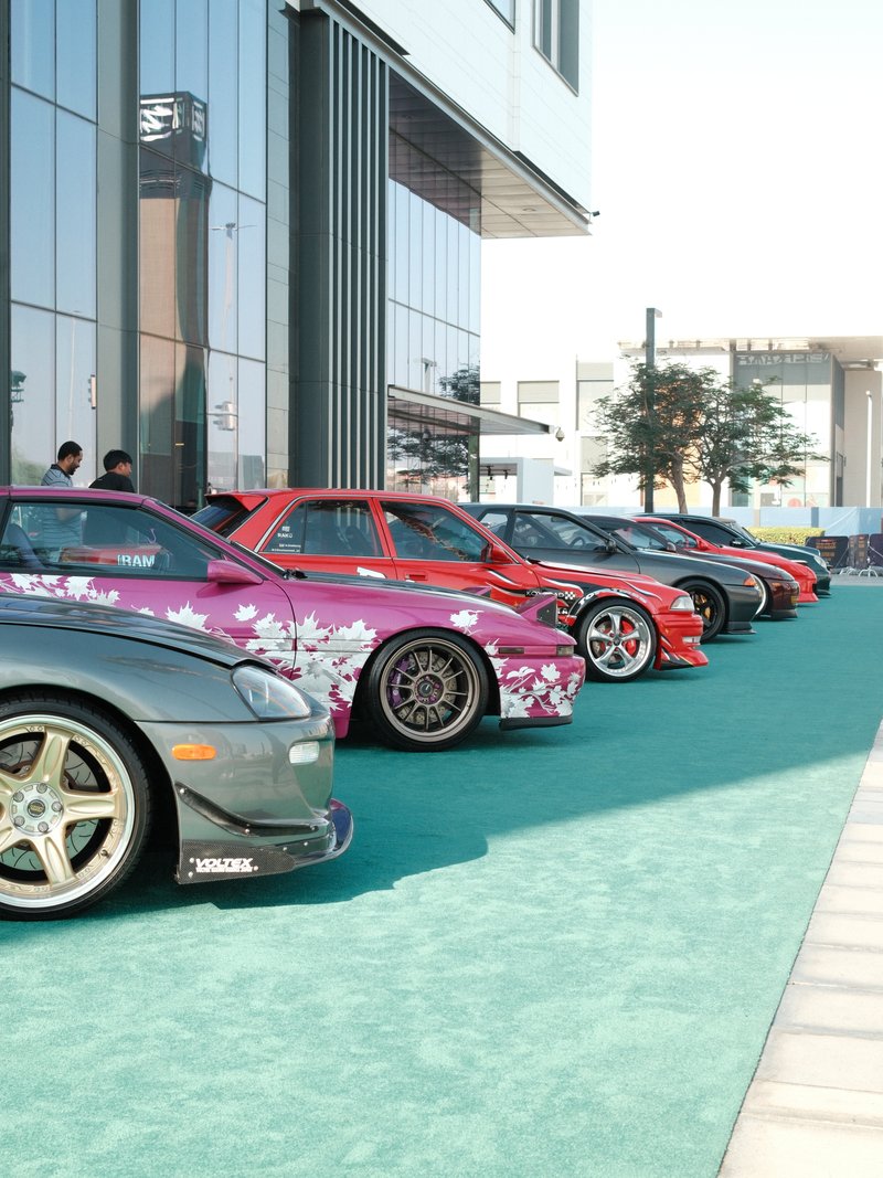 A row of modified cars with floral designs parked on a turquoise carpet outside a modern building.