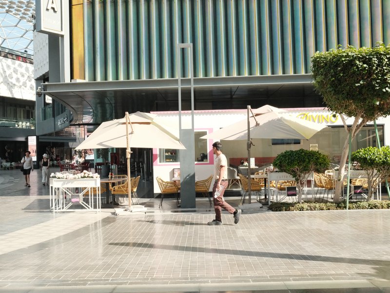 A street scene featuring outdoor dining with umbrellas and a person walking by in a modern urban setting.