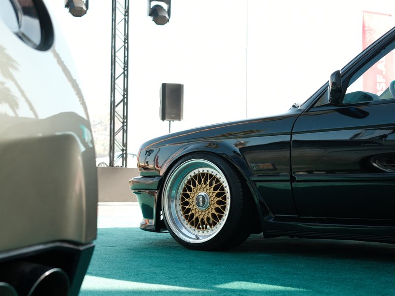 Close-up of two cars at an event, focusing on a black car with gold wheels and a blurred foreground.
