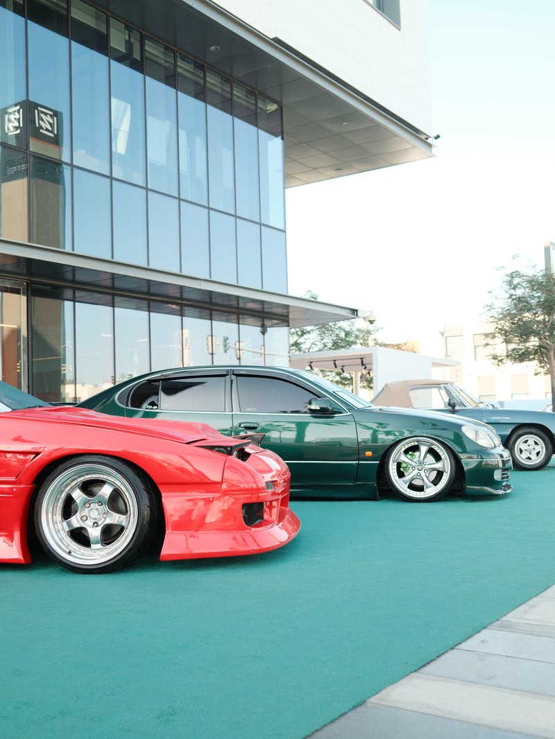 Three modified cars parked on a green surface in front of a modern building.