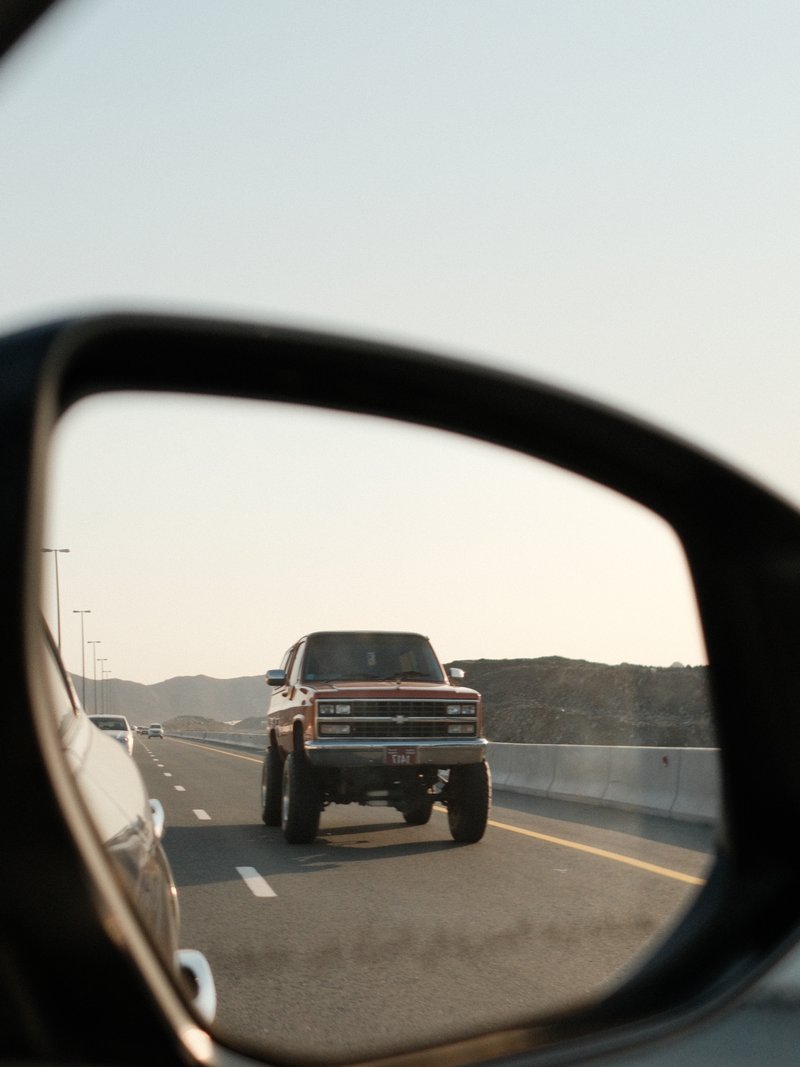 A reflection of a large truck in a side mirror on a highway with a clear sky.