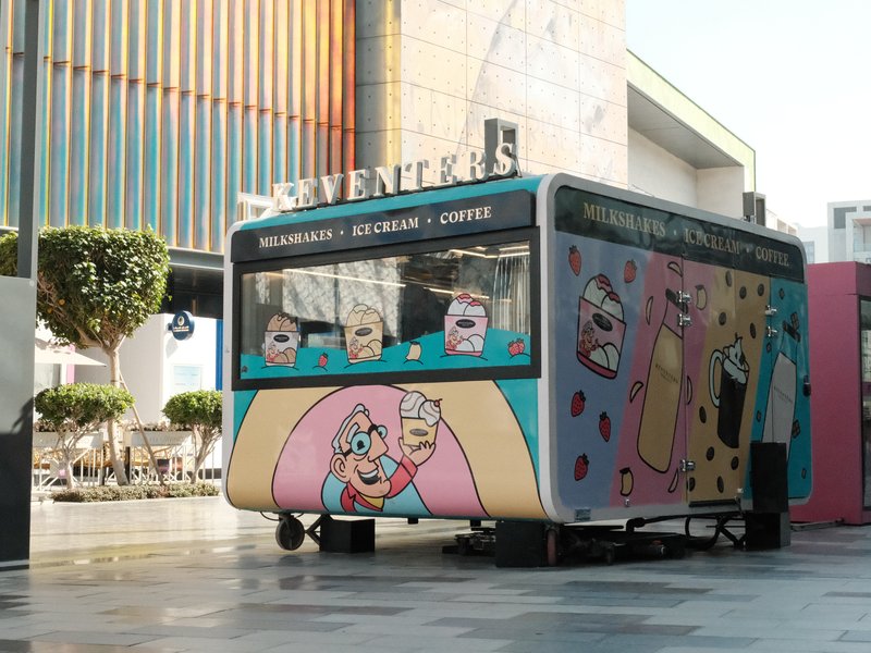A colorful food truck with cartoon characters, parked in an urban plaza with modern architecture in the background.