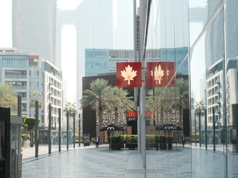 A modern urban scene featuring palm trees and reflective glass buildings with red maple leaf banners.