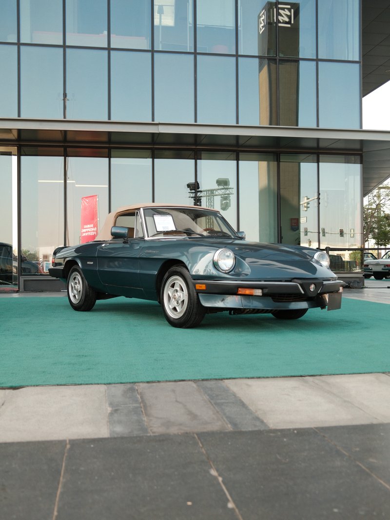 A vintage convertible car parked in front of a modern glass building on a green carpet.