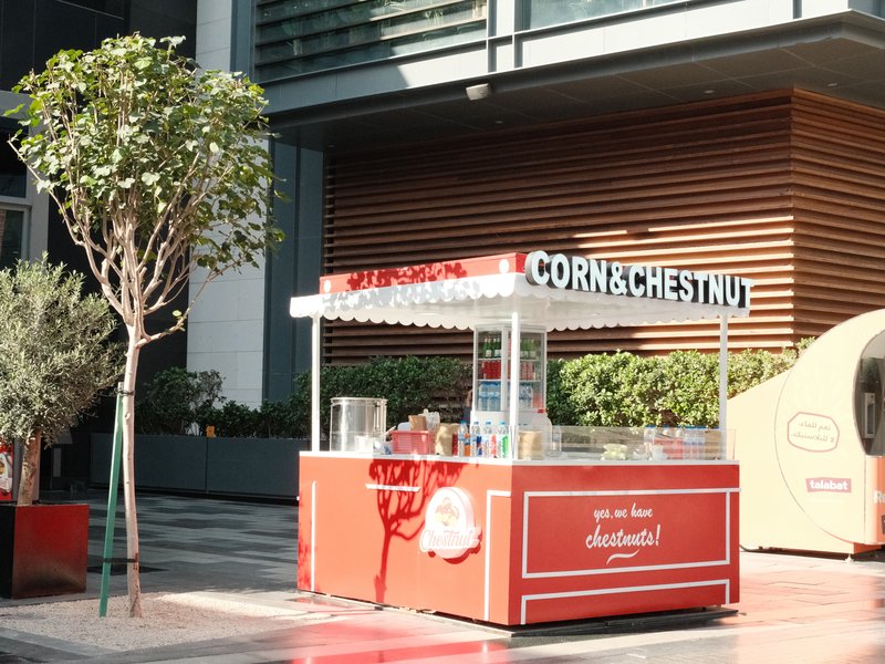 A vibrant red corn vendor cart in an urban setting with greenery and modern architecture in the background.
