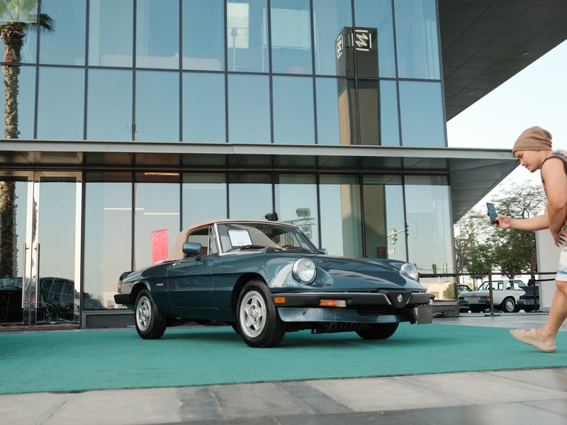 A vintage car parked in front of a modern glass building with a child walking by.