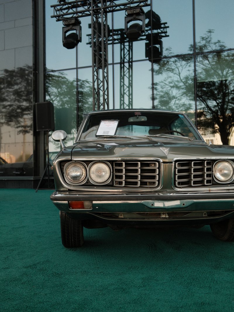 A classic car parked on a green carpet in front of a modern building with reflections and stage lights.