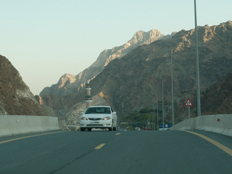 A white car driving on a winding road surrounded by rocky mountains under a clear sky.