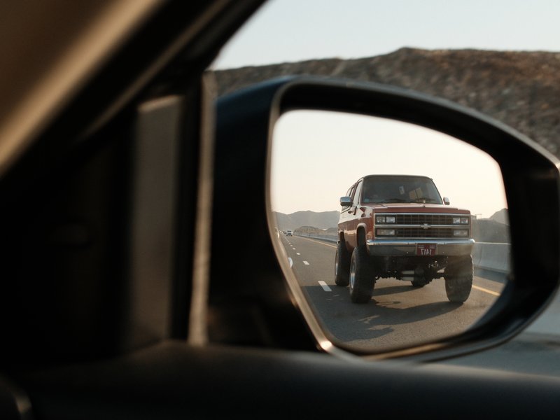 A side mirror view of a large truck driving on a highway with mountains in the background.