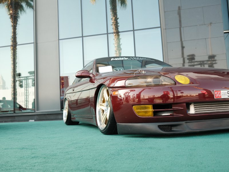 A low-profile maroon sports car parked on a teal carpet with palm trees and a modern building in the background.