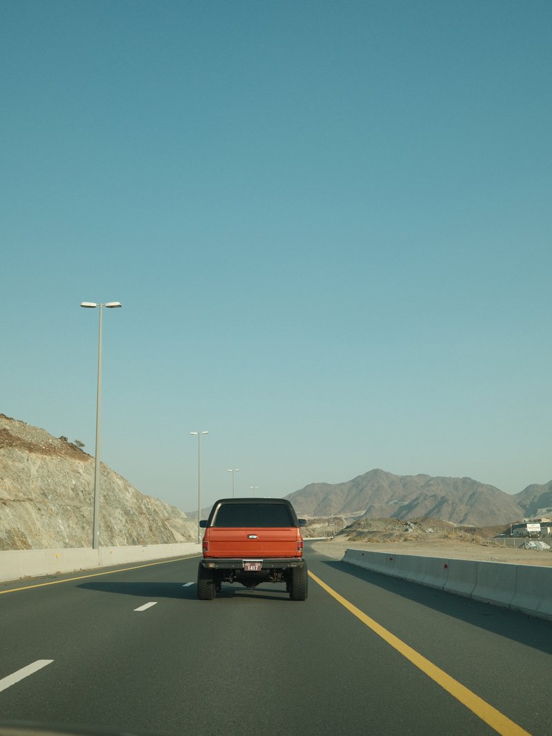 An orange truck driving on a highway with mountains in the background under a clear blue sky.