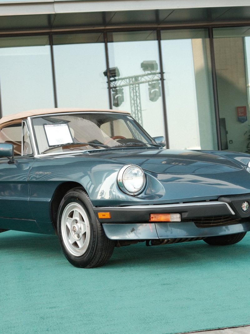 A classic blue convertible car parked indoors on a green carpet with large windows in the background.