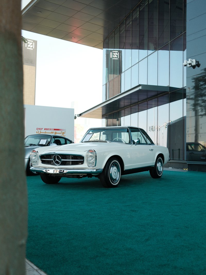 A classic white Mercedes-Benz parked in front of a modern glass building on green turf.