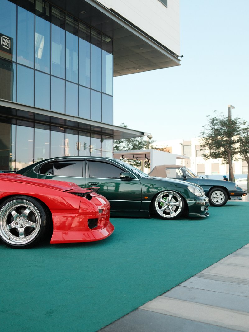 A row of modified cars parked on a green carpet outside a modern building.