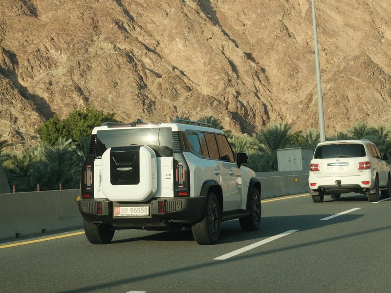 Two vehicles on a highway with rocky mountains in the background under clear skies.