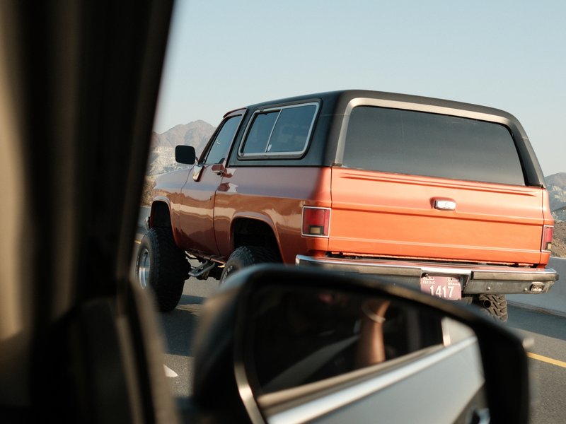 An orange vintage truck driving on a highway, seen from the side mirror of another vehicle.